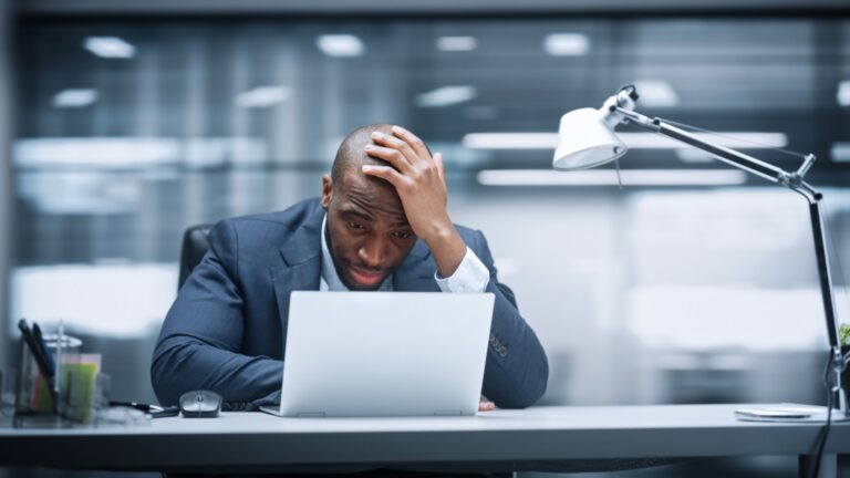 A businessman holding his head while staring at a laptop in an office